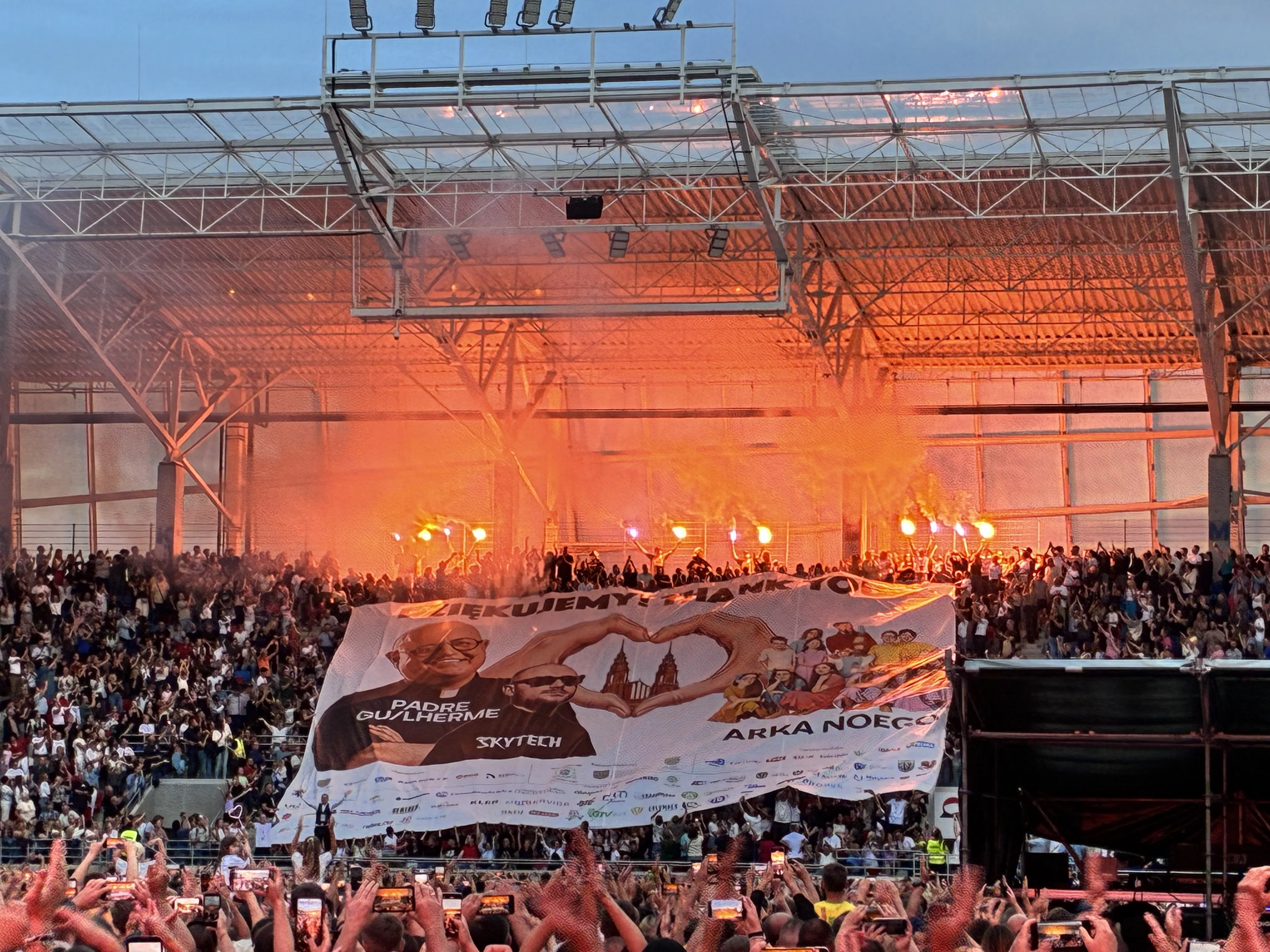 Crowd celebrating at Itaka Arena's first concert, featuring flares and a large banner for performers Padre Guilherme and Skytech.