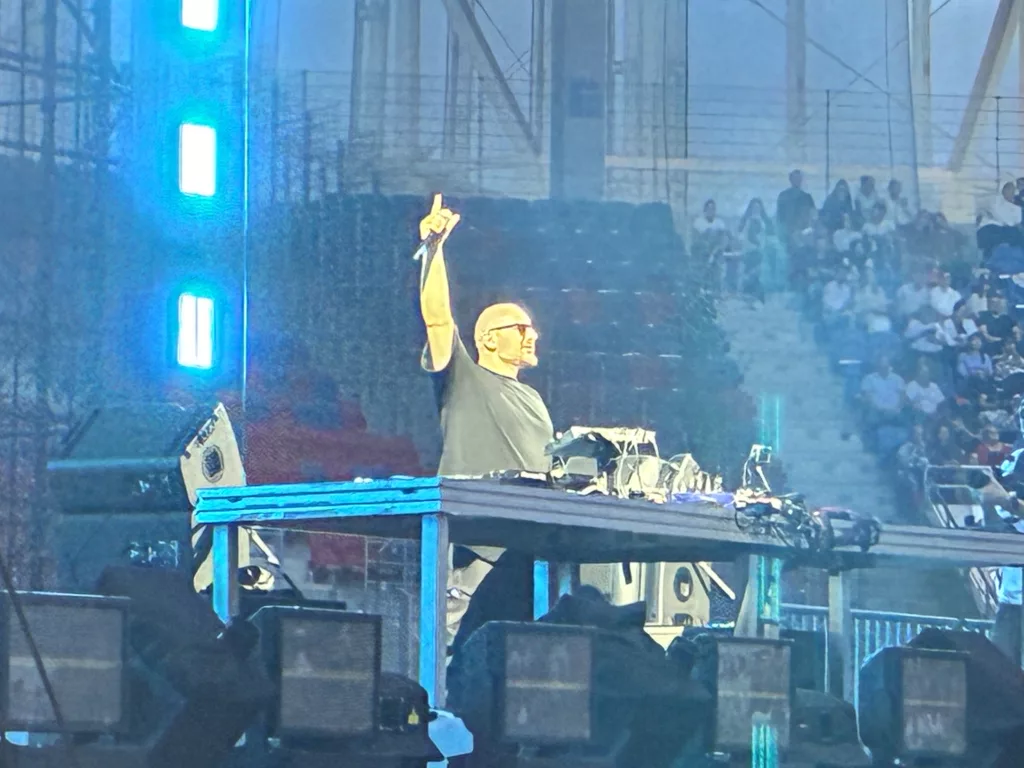 A DJ performs energetically on stage at the inaugural concert of Itaka Arena, surrounded by vibrant lights and an audience.