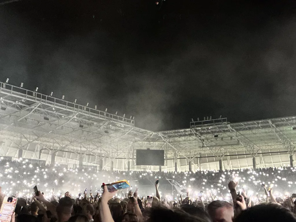Crowd with raised hands and phone lights illuminated in a smoky Itaka Arena during its first concert event.