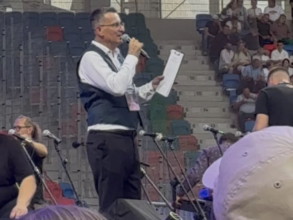 A host speaks on stage at the inaugural concert at Itaka Arena, with musicians and an audience in the background.